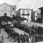 Messa in piazza con podestà inginocchiato e militari schierati. Sulla destra, parzialmente in ombra e indicati dalla freccia, i locali che ospitavano la biblioteca dei confinati (CSDU)