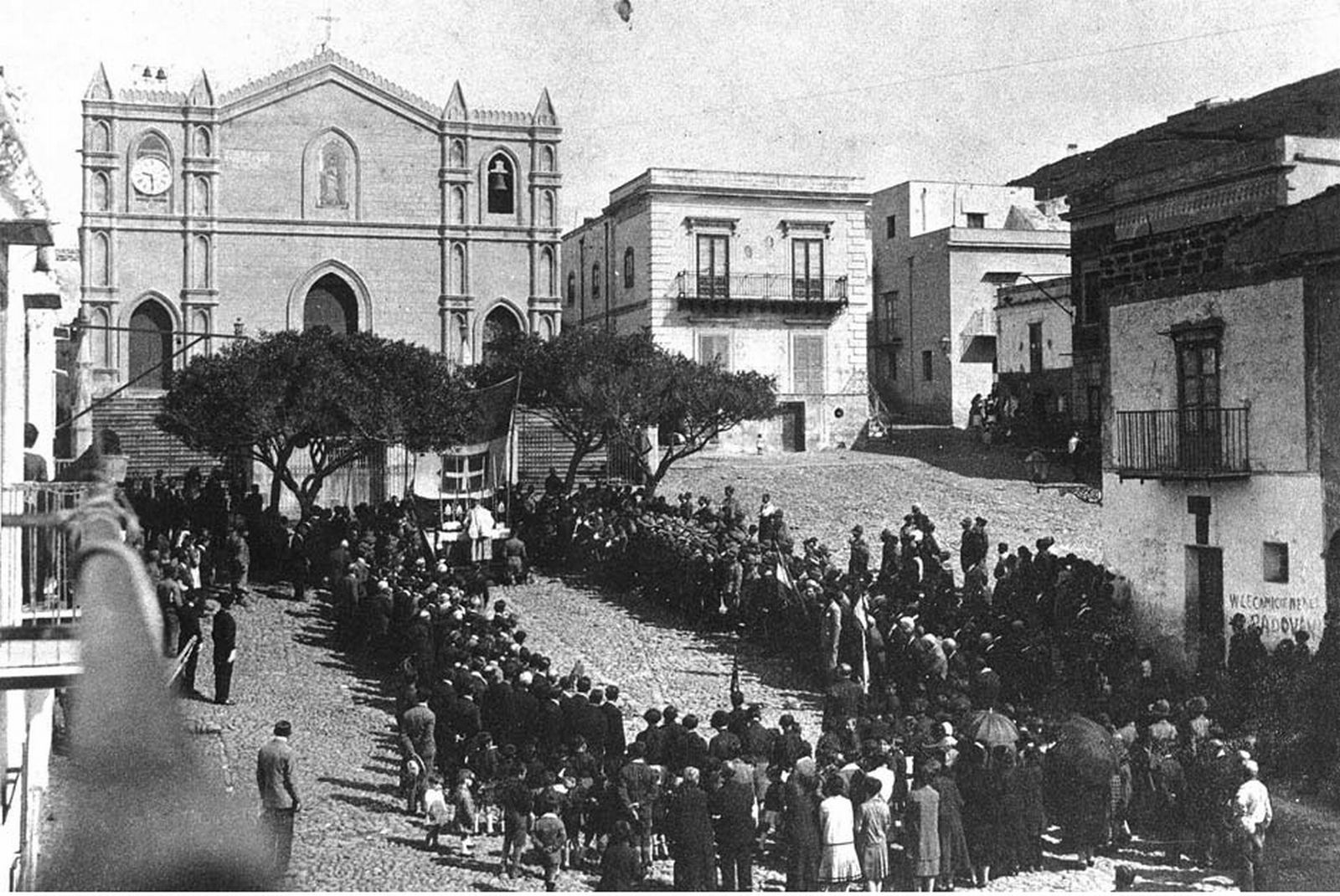 Messa in piazza con podestà inginocchiato e militari schierati. Sulla destra, parzialmente in ombra e indicati dalla freccia, i locali che ospitavano la biblioteca dei confinati (CSDU)