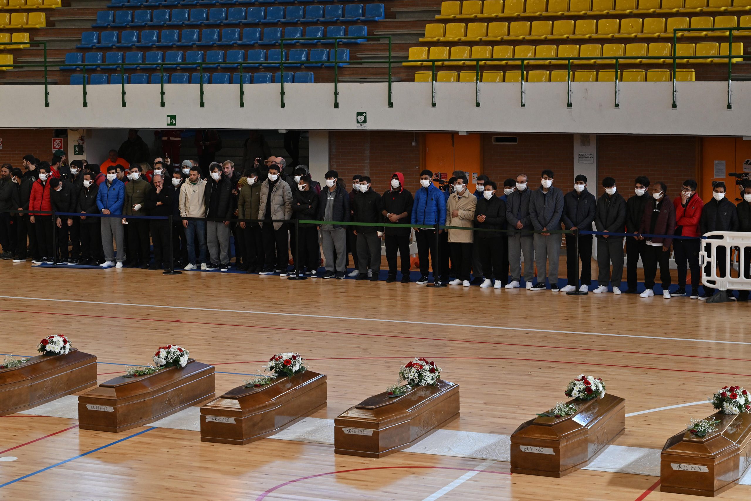 Un nastro separa la vita e la morte, da una parte le persone in visita alla camera ardente, dall'altra le bare delle vittime