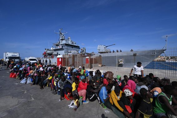 Centinaia di migranti aspettano in fila al molo del porto per essere imbarcati sulla nave militare Cassiopea