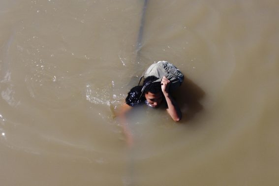 Un ragazzo cerca di attraversare il fiume aiutandosi con una corda, portando il suo zaino sulla testa